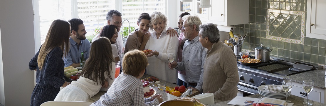 A group of 11 people gathers around a kitchen counter topped with serving plates full of food. 